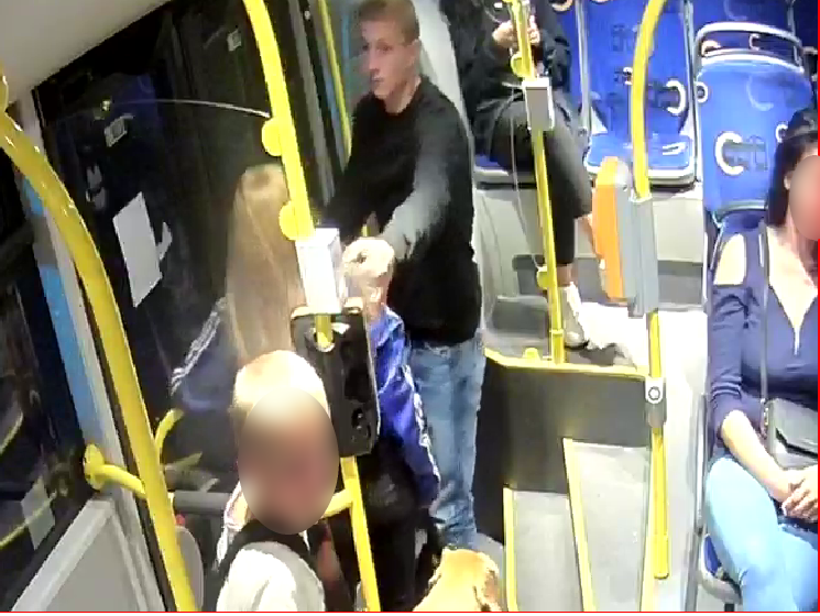 Zdjęcie przedstawia mężczyznę w czarnej bluzie i jasnych spodniach, który znajduje się w autobusie miejskim w towarzystwie pasażerów autobusu. 