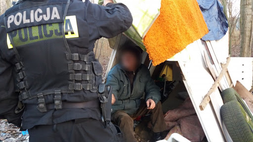 zdjęcie kolorowe: umundurowany policjant podczas rozmowy z bezdomnym mężczyzną mieszkającym w szałasie w lesie