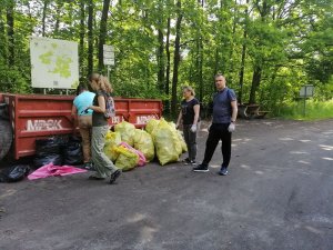 Na zdjęciu widać jak dzielnicowy stoi obok kontenerów na śmieci obok uczestnicy sprzątania lasu wrzucają śmieci do kontenerów