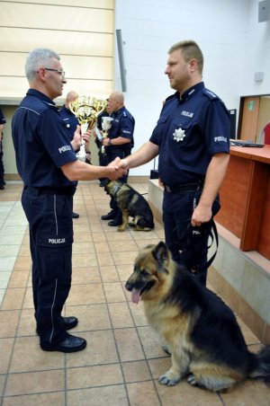 Na zdjęciu widać jak oficer policji wręcza puchar i dyplom przewodnikowi psa który stoi ze swoim psem
