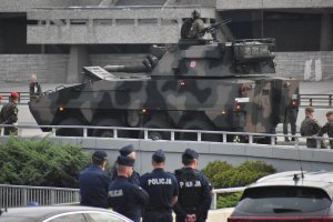 Na zdjęciu widać policjantów stojących na rondzie przed nimi jedzie transporter opancerzony z tyłu widać budynek spodka