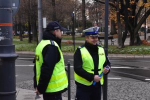 Na kolorowym zdjęciu widać dwóch policjantów ruchu drogowego obok przejścia dla pieszych jak stoją trzymając elementy odblaskowe, w tle widać plac wolności w Katowicach