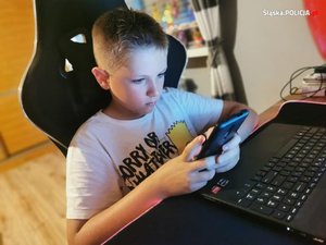 zdjęcie kolorowe: chłopiec siedzący przed monitorem i korzystający z telefonu komórkowego