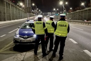 zdjęcie kolorowe: trzech policjantów wydziału ruchu drogowego stojących kolo radiowozu