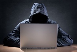 zdjęcie kolorowe: mężczyzna ubrany w ciemną bluzę z założonym kapturem na głowie siedzący przed laptopem